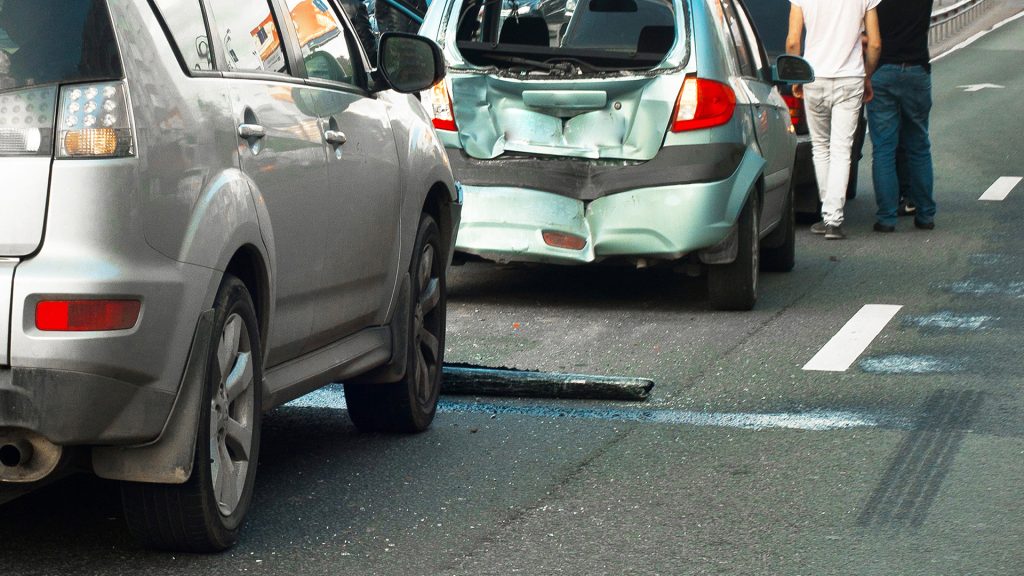 motor-vehicle-accident-lawyer-north-carolina Daytime scene of a two-vehicle collision on a suburban road, showing a silver sedan with a crumpled front end and a gray SUV with rear-end damage, surrounded by scattered debris and a background of trees and power lines.